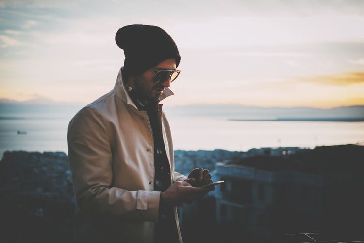Man using phone while standing against city during sunset