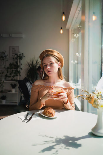 French female in beret sitting at table in cafe with aromatic glass of coffee and freshly baked croissant