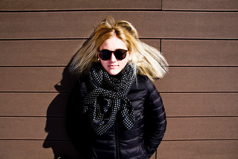 Portrait of young woman wearing sunglasses and warm clothing while standing against wall