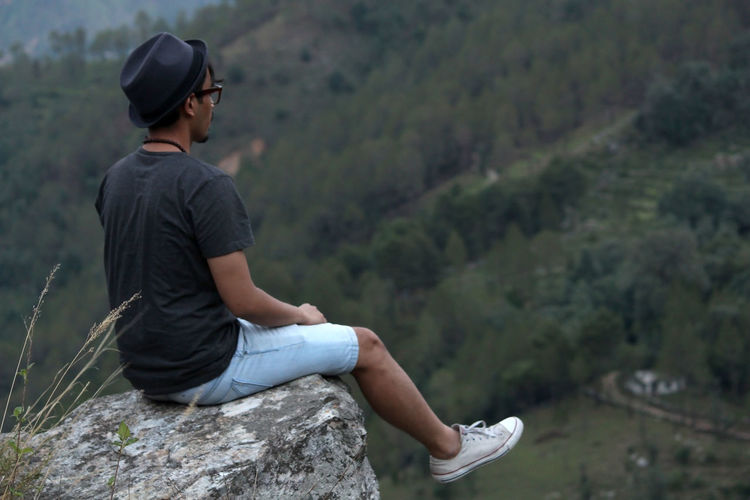 Young man wearing a cool hat sitting on the edge of a rock in front of a mountain range.