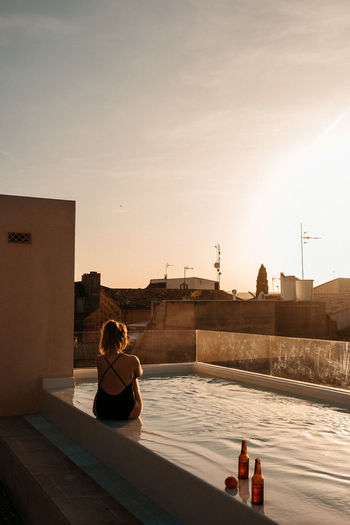 Back view of unrecognizable young female traveler with long hair in stylish black swimsuit sitting in swimming pool and admiring sunset over city during summer holidays in mallorca