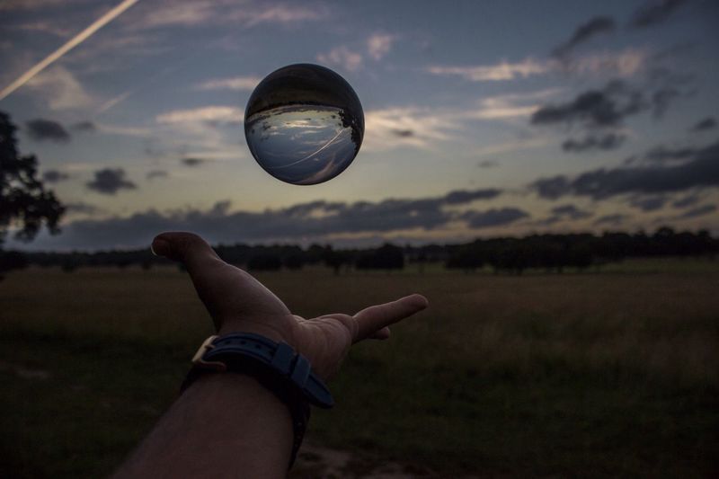 Cropped hand of man holding crystal ball in mid-air against sky during sunset