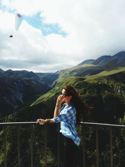 Woman standing by railing with person paragliding against cloudy sky