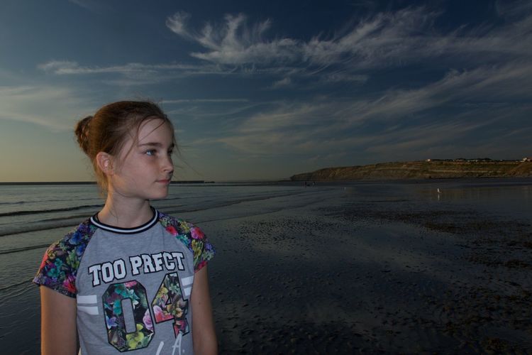 Girl looking away while standing at beach against sky during sunset