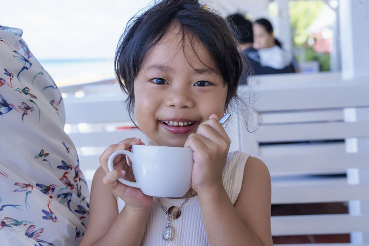 Close up of adorable little girl holding a white cup of hot drink and smiling at the camera. 