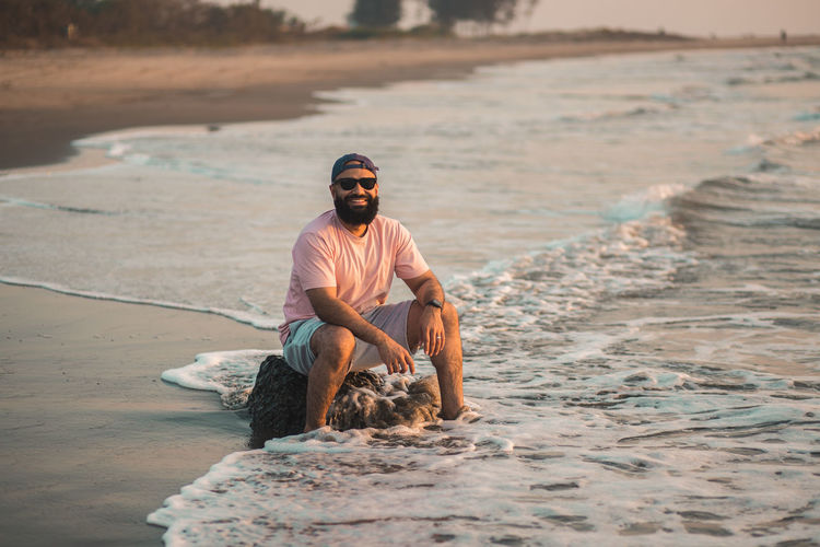 Full length of good looking bearded man sitting at the beach during sunset with feet in the water