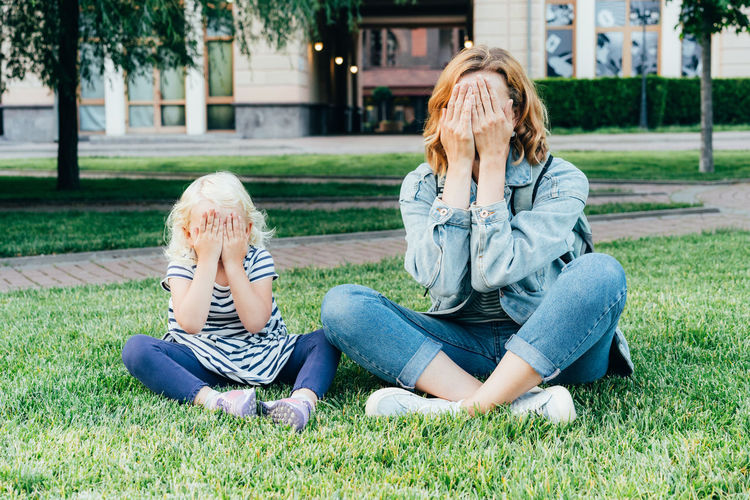 Mom and her little daughter sitting on the lawn covered their faces with hands playing hide and seek