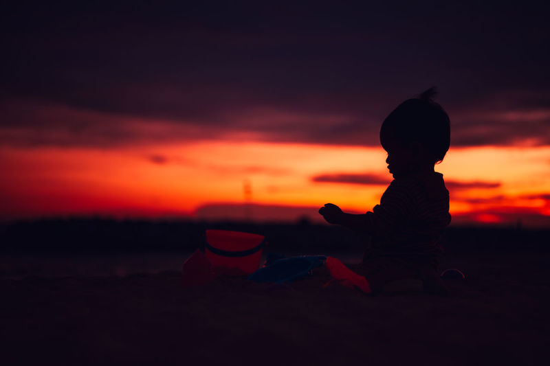 Silhouette boy sitting on land against sky during sunset