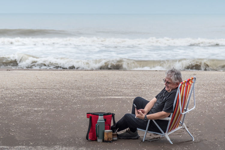 Mature man with gray hair sitting on a beach chair smiling while looking at his smartphone.