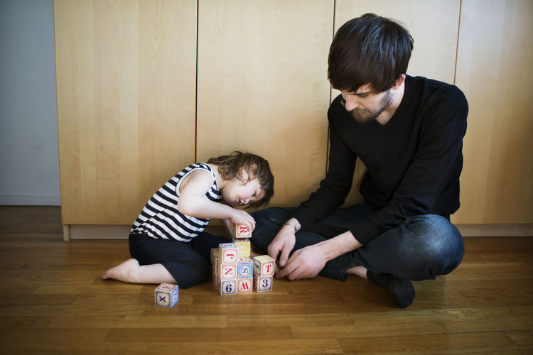 Father looking at daughter playing with toys blocks while sitting on floor