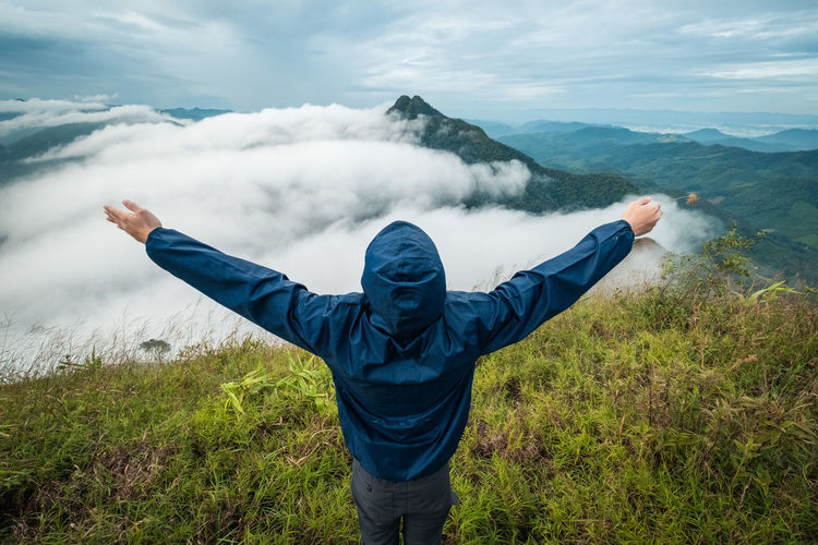 Rear view of person with arms outstretched looking at landscape against cloudy sky