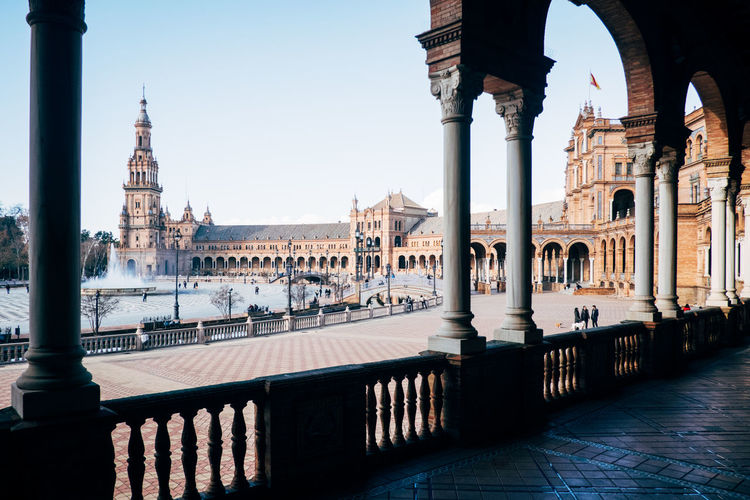 Historic buildings at plaza de espana seen through colonnade against blue sky