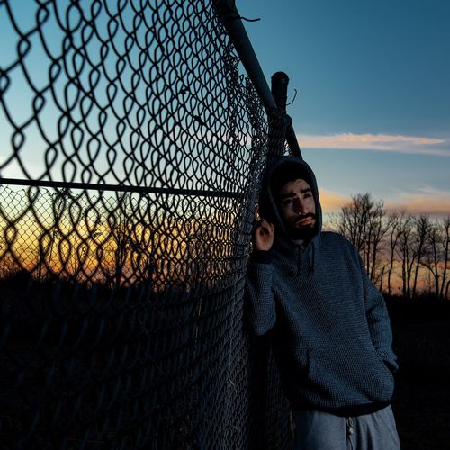 Thoughtful young man standing by fence against sky during sunset