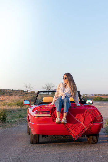 Vertical view of young teenager sitting relaxed on a red convertible vintage car enjoying the summer