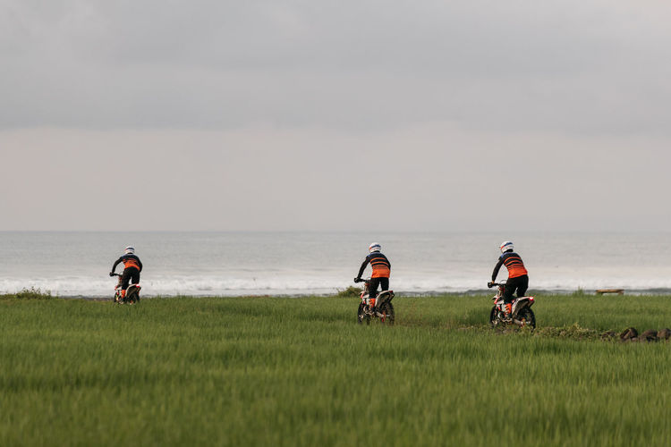 People riding dirt bikes on grass by sea against sky