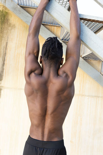 Shirtless fit man hanging from staircase stretching his back