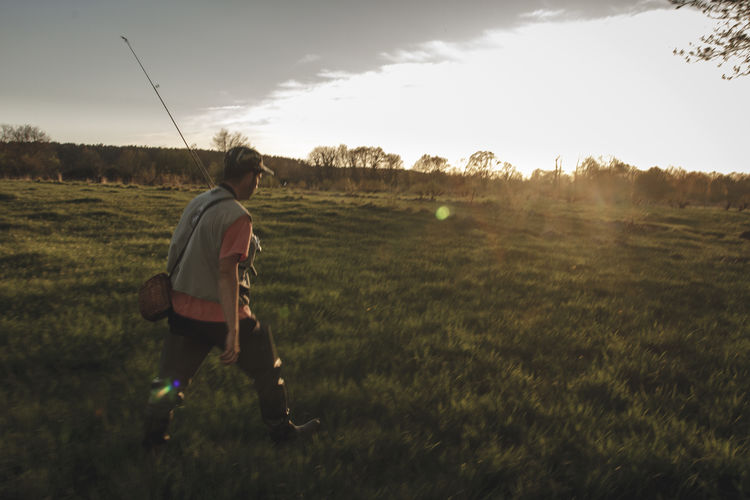 Full length side view of man with fishing rod walking on field against sky during sunset