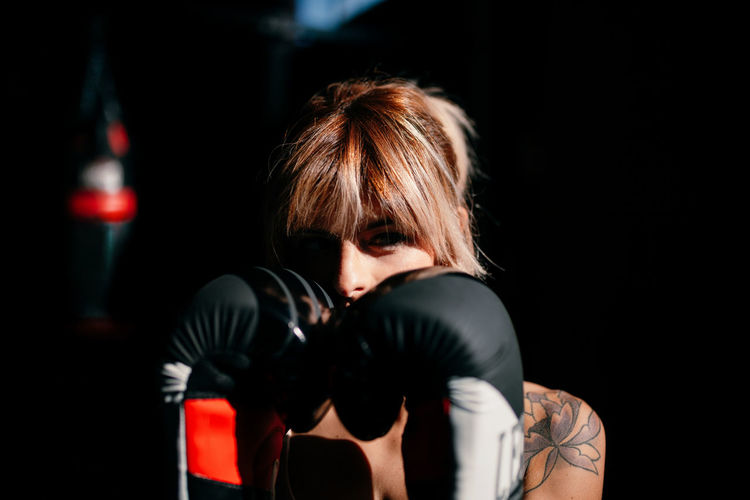 Portrait of young confident female athlete looking at camera in boxing gloves standing in defensive stance during training in dark gym