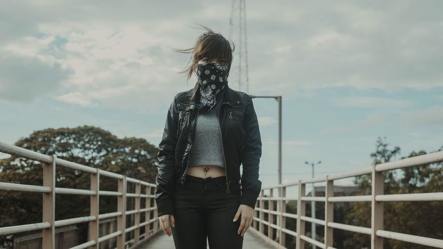 Woman covering face with handkerchief while standing on footbridge against sky