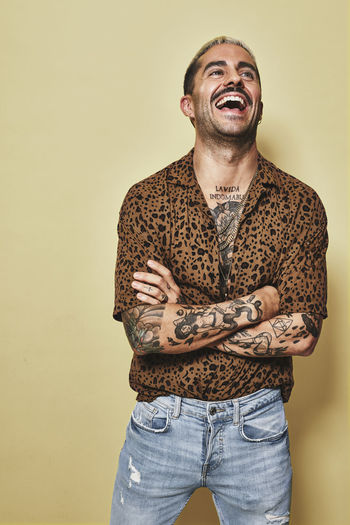 Cheerful fashionable male model with tattoos wearing trendy leopard shirt and jeans standing against beige background and looking away