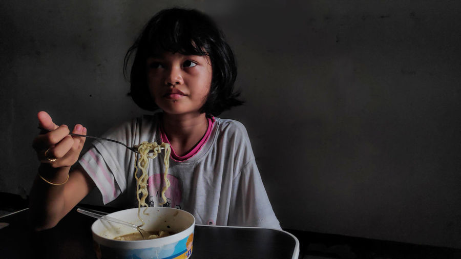 Cute asian child girl eating delicious instant noodles with fork in the dark background