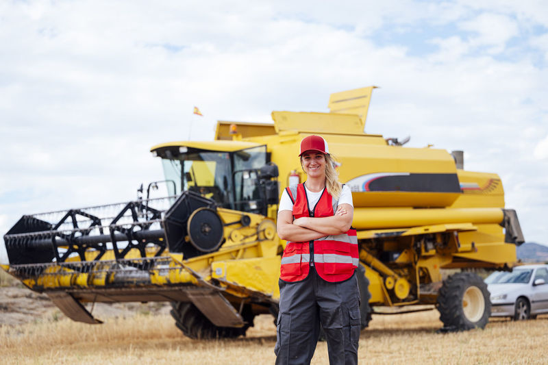 Positive female worker in uniform looking at camera while standing in agricultural field with industrial combine harvester in rural area