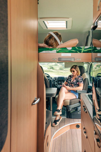 Woman listening music lying on camper van bunk bed while her friend looking cellphone