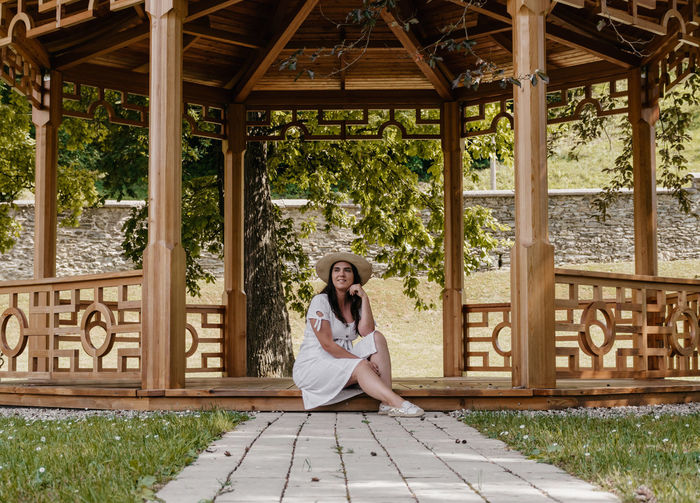 Portrait of beautiful young woman wearing white dress sitting under wooden pavilion in park.
