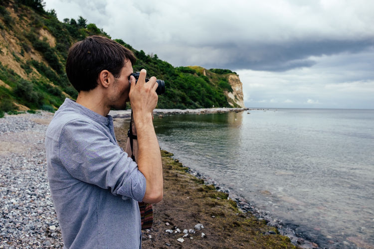 Side view of man photographing sea with camera at beach against cloudy sky