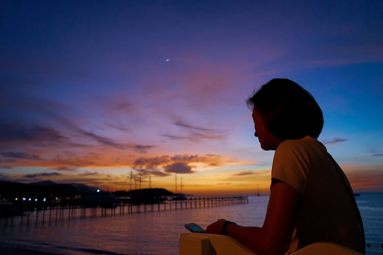Woman using phone at beach against sky during sunset