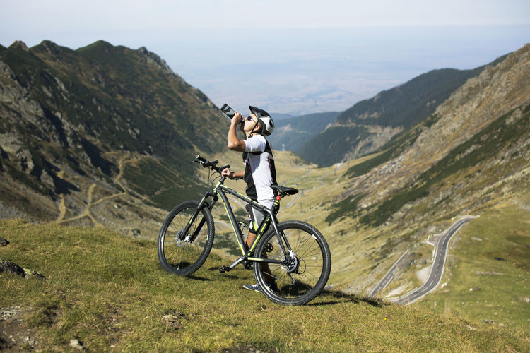 Side view of athlete drinking water while standing by bicycle on mountain