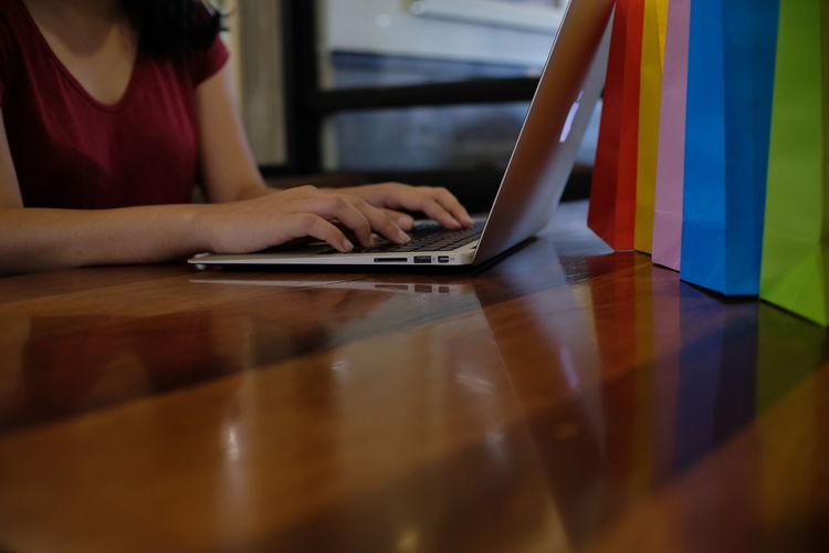 Close-up of woman hands using laptop on wooden table by colorful shopping bags