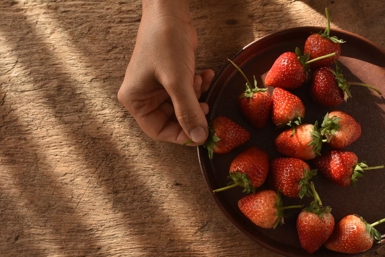 Hand of asian lady picking up fresh strawberry from ceramic plate on wooden table.