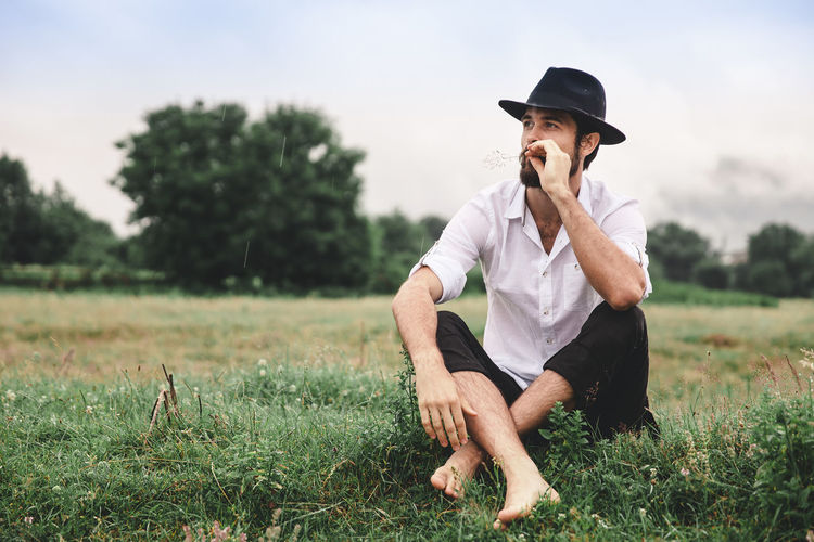 Full length of thoughtful young man looking away while sitting on grassy field