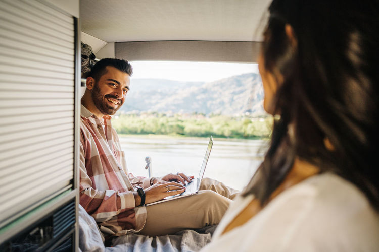 Content hispanic man working on laptop while sitting in camper during trip and looking at unrecognizable woman