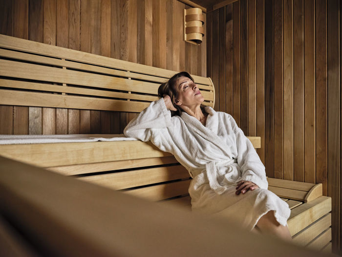 Senior woman with hands behind head relaxing in sauna at health spa
