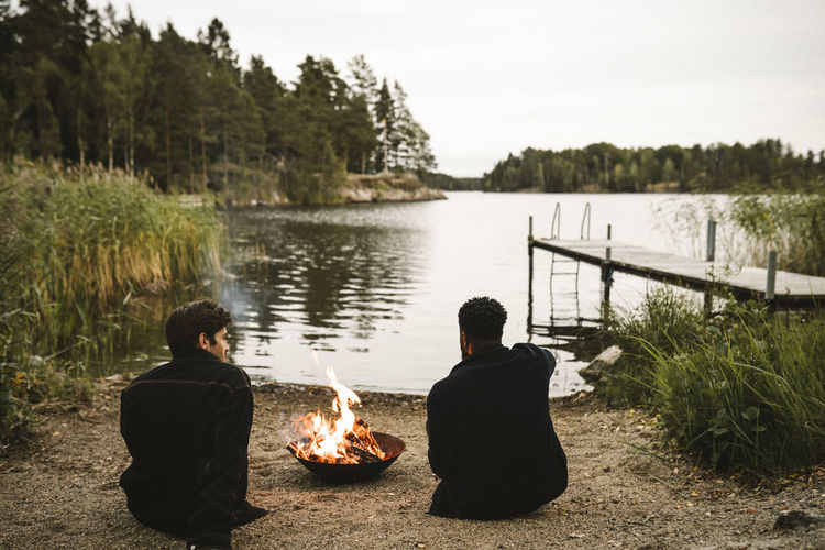 Rear view of male friends talking by fire pit against lake