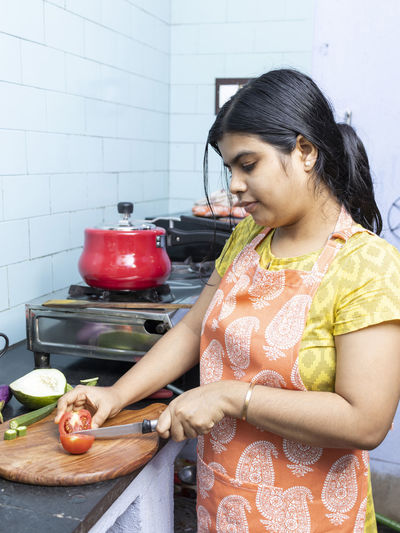 A pretty indian young woman wearing apron cooking and cutting vegetables in domestic kitchen