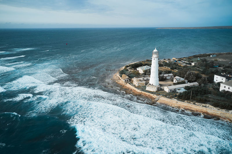 From above drone view of white lighthouse tower and small settlement located on rocky coast washed by foamy sea waves