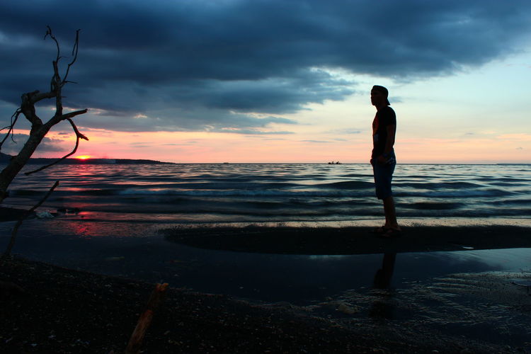 Silhouette man standing at beach against cloudy sky during sunset