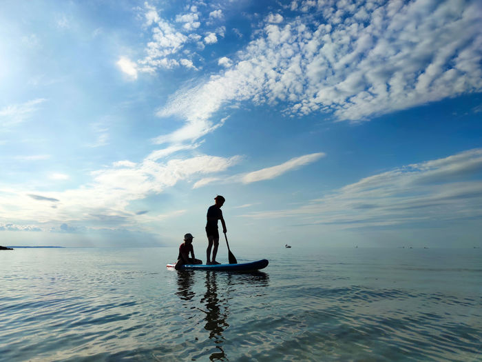 Silhouette siblings paddleboarding on sea against sky