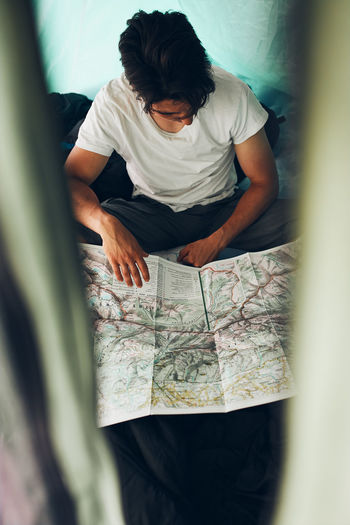 Young man looking at a map of mountain trails planning next trip on summer vacation sitting in tent
