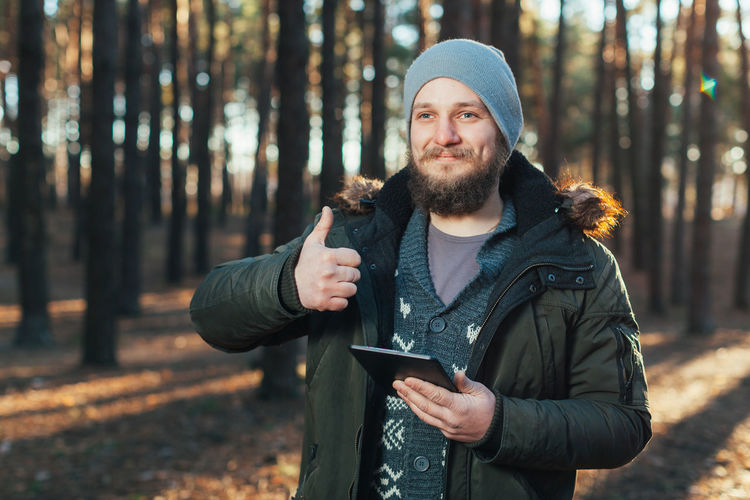 Smiling man showing thumbs up while holding digital tablet in forest