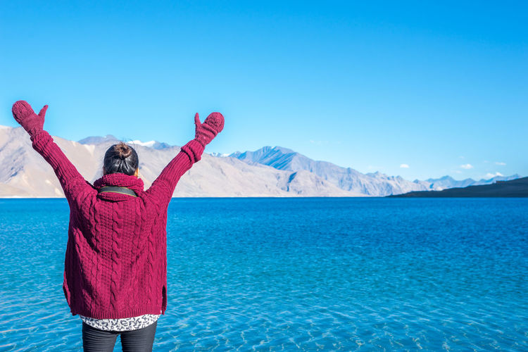 Woman standing by lake with arms raised against clear blue sky