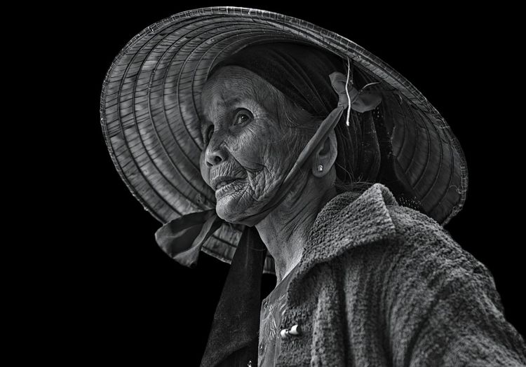 Thoughtful senior woman wearing asian style conical hat against black background