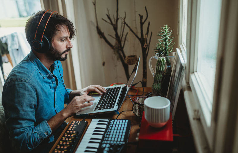 Side view of young man in headphones using synthesizer and laptop at table at home
