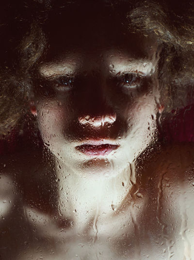 Close-up portrait of young woman seen through wet glass