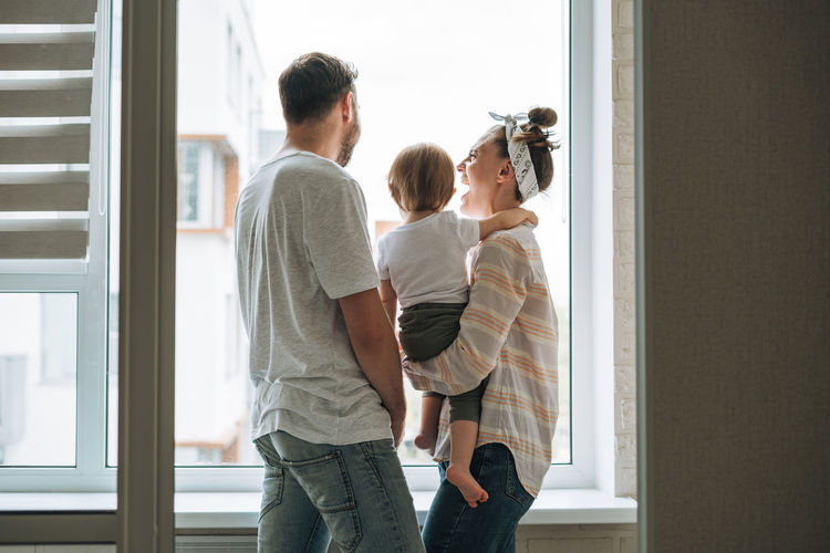 Young family man father and mother woman with baby girl on window sill looking at window at home