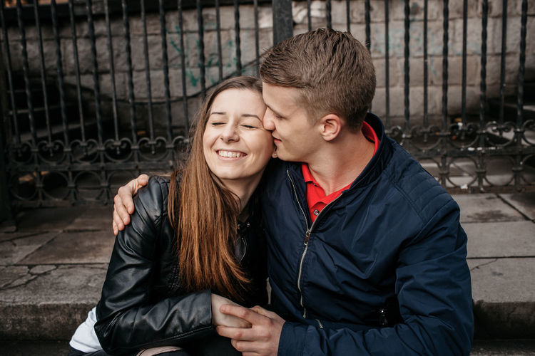 Close-up of boyfriend kissing girlfriend while sitting against gate in city