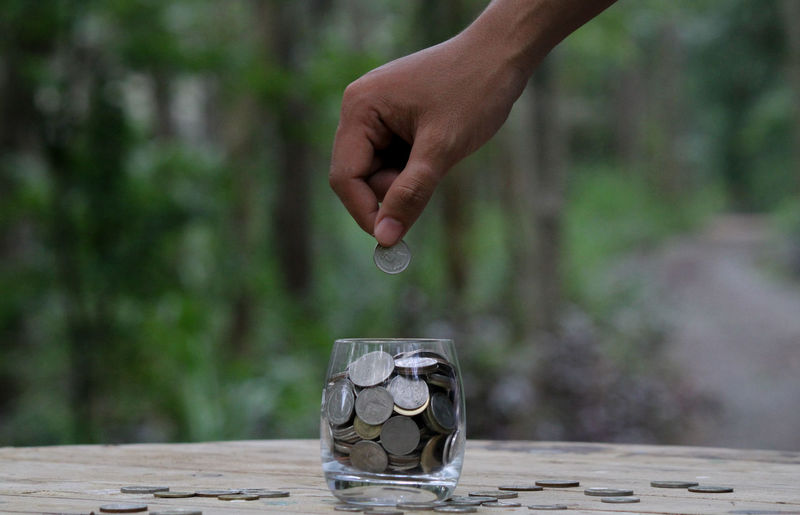 Cropped hand of man dropping coins in drinking glass on table against trees